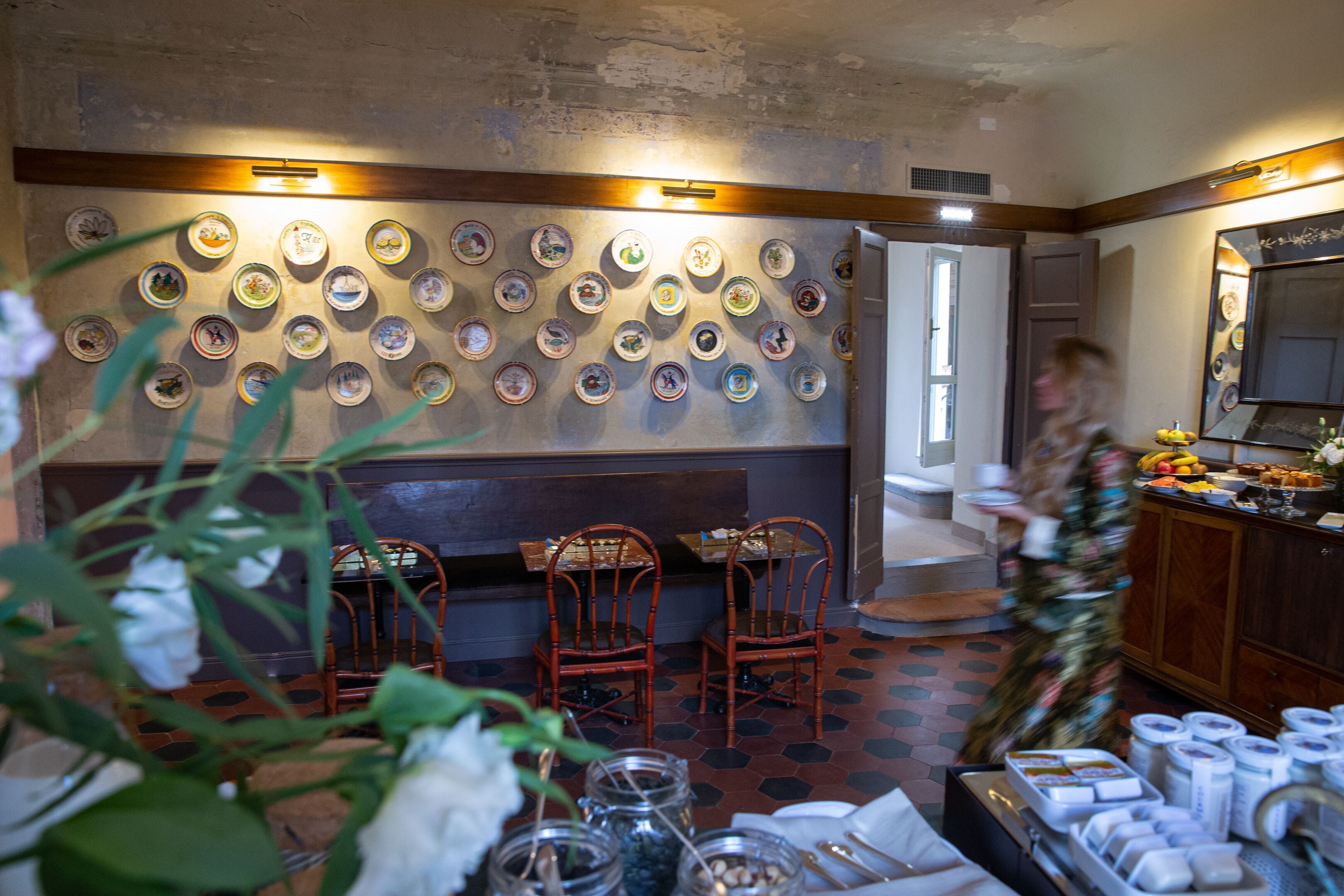 A restaurant interior with a wall of decorative plates, wooden chairs, and a buffet area with food items.
