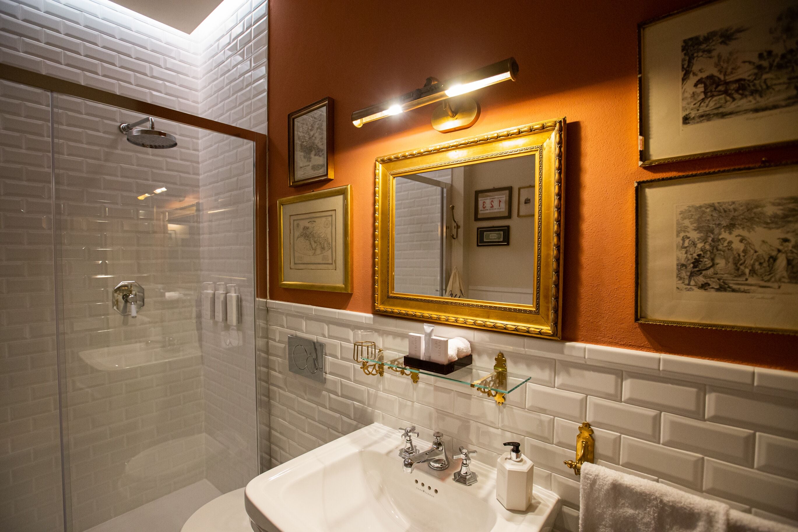 Un baño con ducha envuelta en vidrio, un lavamanos blanco con detalles dorados y obras de arte enmarcadas en las paredes.