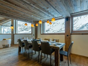 A dining area with a wooden table set for six, surrounded by chairs, with large windows offering a view of a snowy mountain. - Luxurious 5-Bedroom Chalet Apartment with Panoramic Mountain View, Wi-Fi, and Garage (Val-d'Isère)