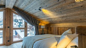 A cozy wooden cabin room with a large bed, a window with a view of snowy mountains, and a lamp on the wall.