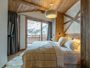 A cozy bedroom with a wooden ceiling, a large bed with a plaid blanket, a nightstand, and a window with a view of snow-covered mountains. - Luxurious 5-Bedroom Chalet Apartment with Panoramic Mountain View, Wi-Fi, and Garage (Val-d'Isère)