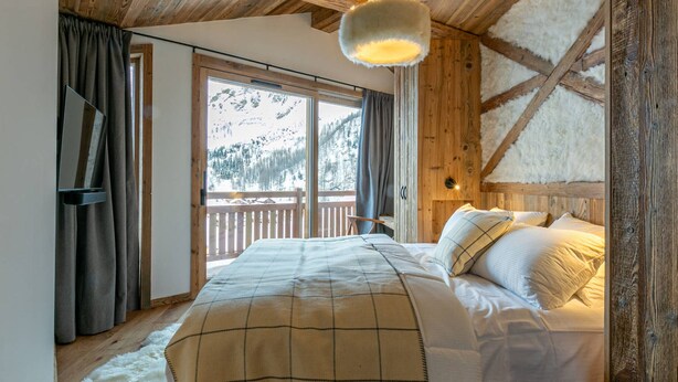 A cozy bedroom with a wooden ceiling, a large bed with a plaid blanket, a nightstand, and a window with a view of snow-covered mountains.