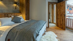 A bedroom with a wooden ceiling, a stone wall, a bed with a patterned blanket, and a view of a building through a wooden door.