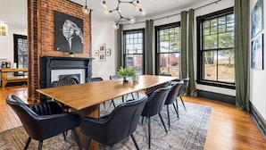 Un comedor con una mesa de madera, sillas negras, chimenea de ladrillo y ventanas amplias con cortinas verdes.
