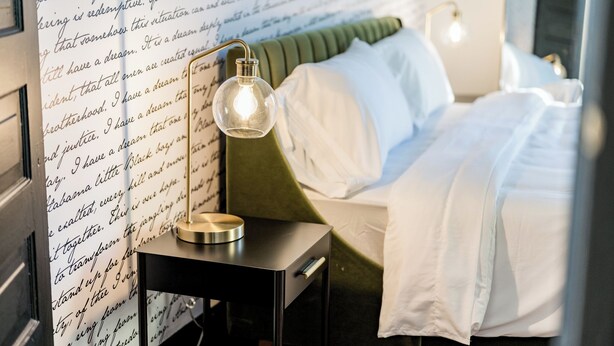 Una cama bien hecha con sábanas blancas, un cabecero tapizado en verde y una mesita de noche con una lámpara.