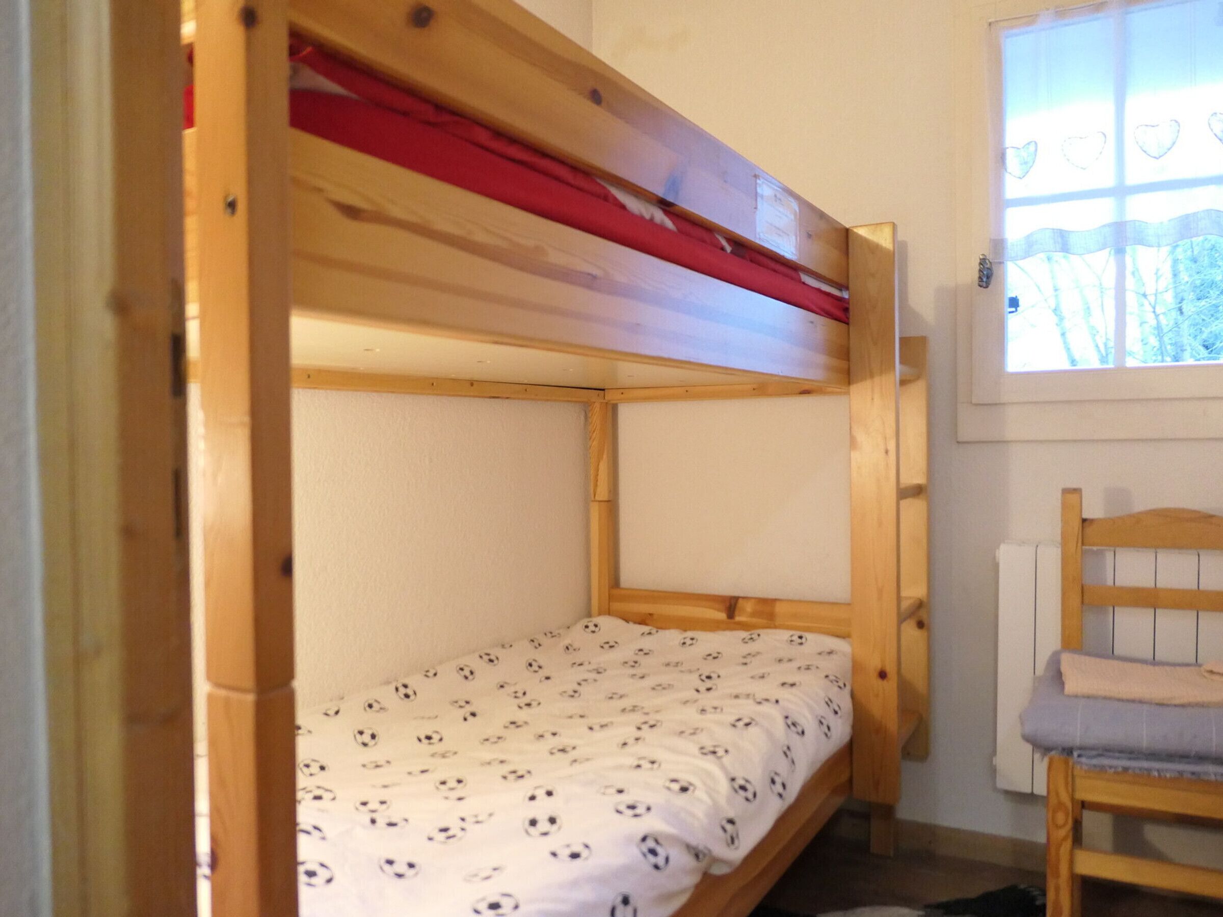 A bunk bed with a red headboard, a wooden chair, and a window with a view of trees.