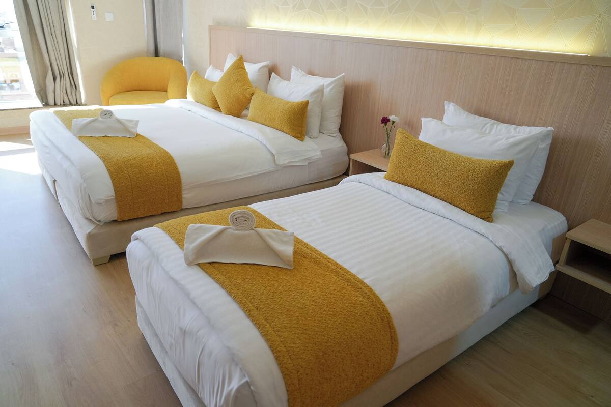 Habitación de hotel con dos camas, cada una con ropa blanca y detalles en amarillo, cabeceros de madera y una mesita de noche con un jarrón de flores.