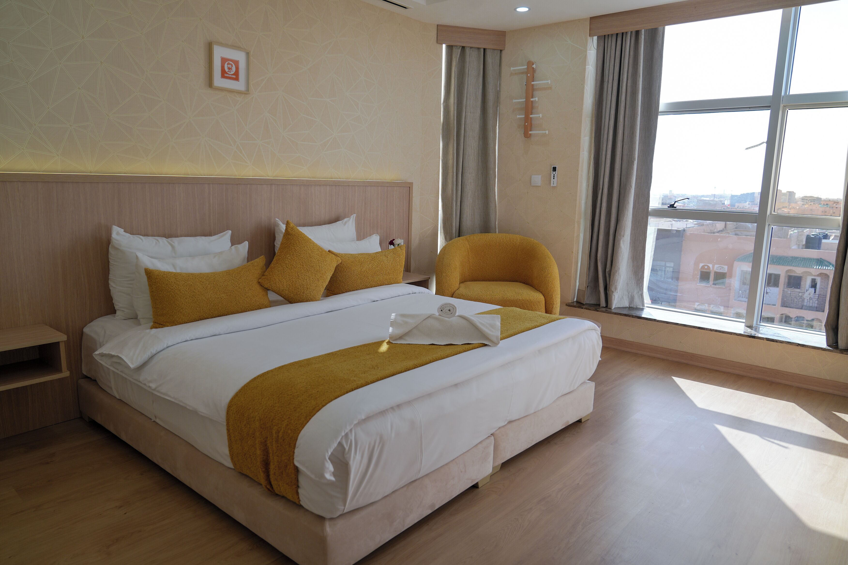 A hotel room with a large bed, a chair, a window with a city view, and a wall-mounted light fixture.