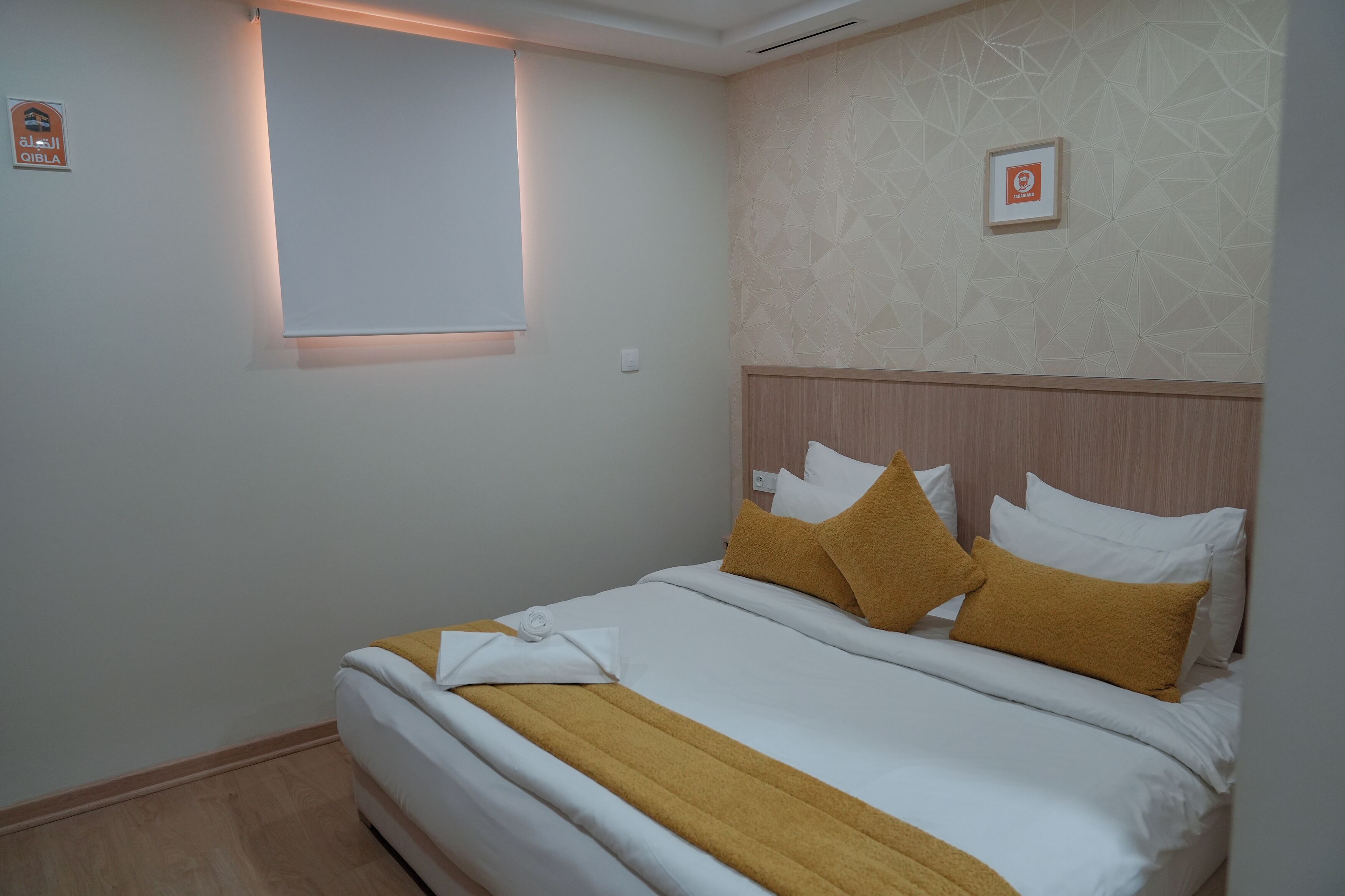 A neatly made bed with white and mustard bedding, a wooden headboard, and a geometric patterned wall.