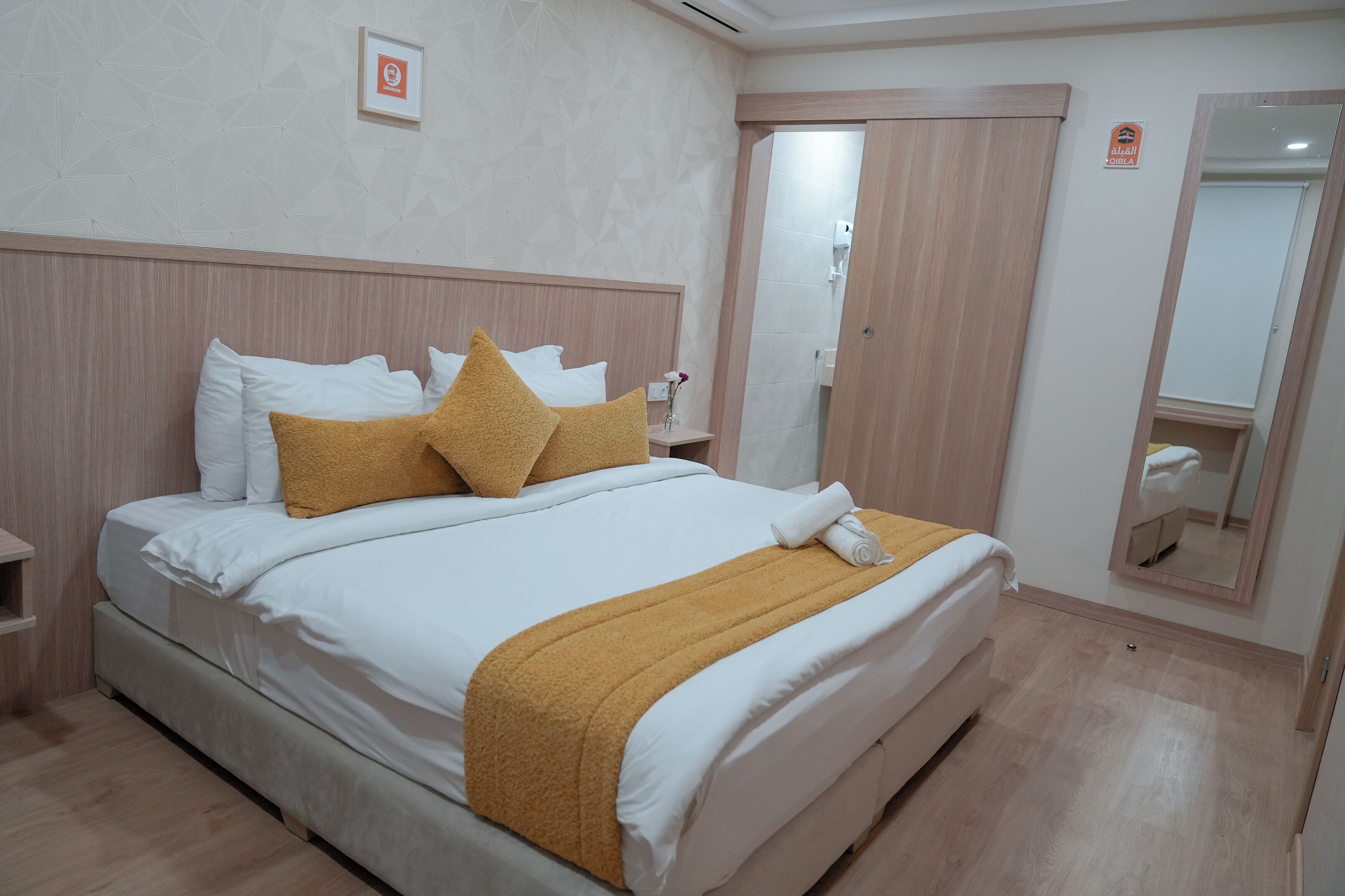 Una cama bien hecha con ropa de cama blanca y mostaza, un cabecero de madera y una mesita de noche con un jarrón.