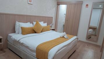 Una cama bien hecha con ropa de cama blanca y mostaza, un cabecero de madera y una mesita de noche con un jarrón.