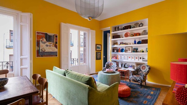 Una habitación con sofá verde, una mesa de comedor, un sillón estampado, una lámpara roja y una estantería llena de objetos diversos.
