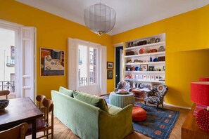 Um quarto com um sofá verde, uma mesa de jantar, uma poltrona estampada, um abajur vermelho e uma estante repleta de objetos diversos.