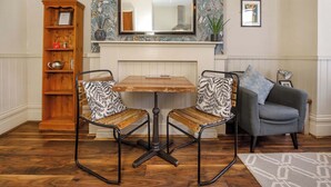 A dining area with a wooden table and two chairs, a bookshelf, a grey sofa, and a mirror above the fireplace.