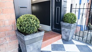 A building entrance with a black door, two square planters with green shrubs, and a checkered floor.