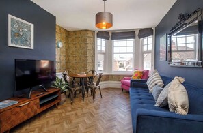 A living room with a TV, wooden herringbone floor, a dining table with chairs, a blue sofa with cushions, and large windows with a view of buildings. - Excellent Two bedroom apartment in central York A3 (York)
