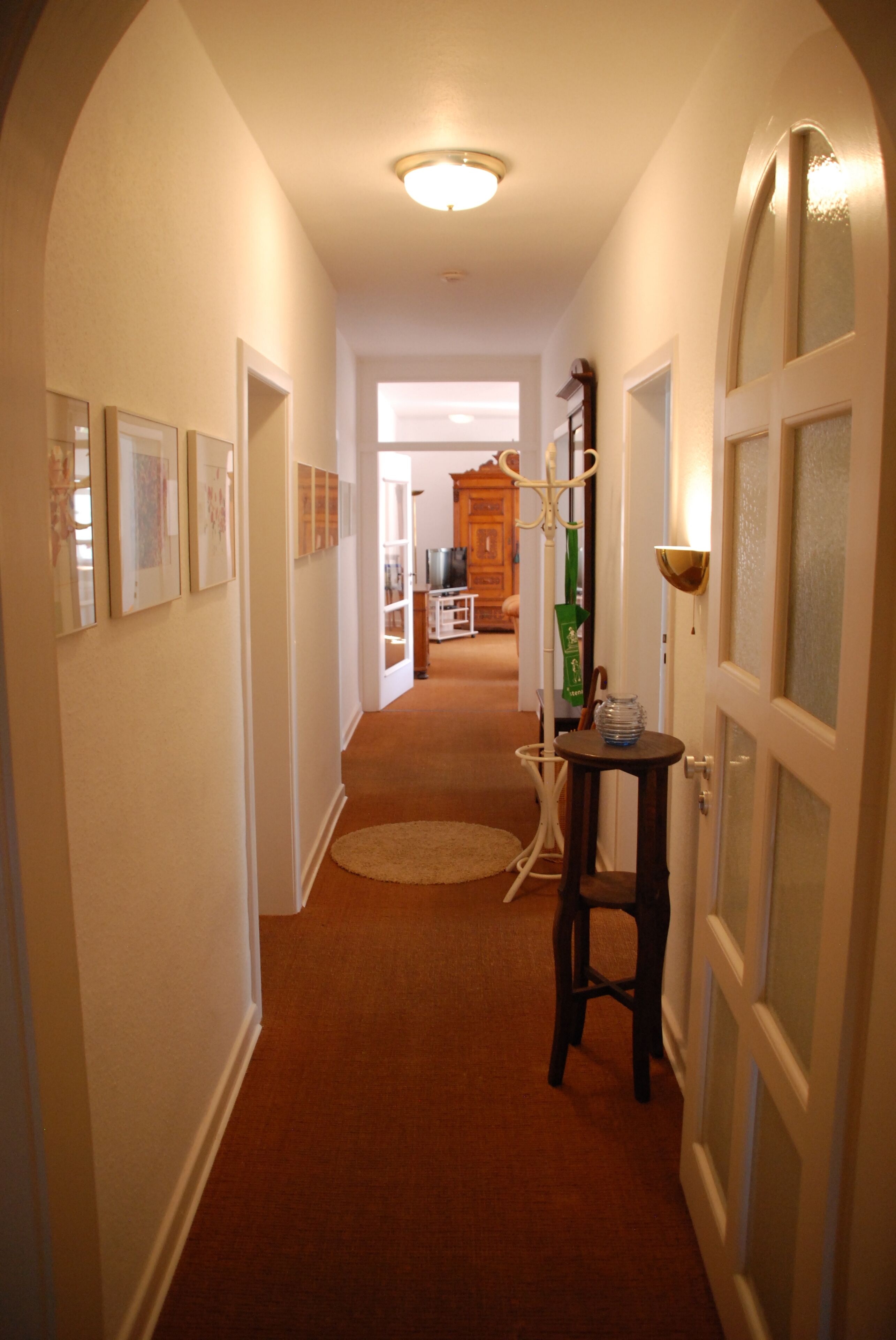 A hallway with a carpeted floor, framed pictures on the walls, a round rug, a small table with a lamp, and a door with frosted glass panels.