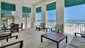 Un balcon spacieux offrant une vue sur l’océan, aménagé avec des tables et des chaises en bois, et de grandes fenêtres dotées de volets verts.