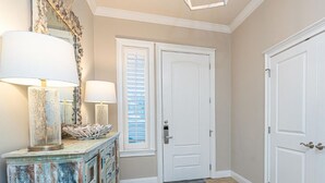 Un couloir avec une porte blanche, un miroir doté d’un cadre décoratif et deux lampes posées sur une commode rustique.