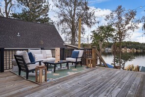A deck with outdoor seating, a wooden table, and a view of a lake with trees and a house in the background.