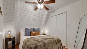 Une chambre avec un ventilateur de plafond installé sous un toit en pente, un lit recouvert d’une couverture grise, deux lampes de chevet et une armoire.