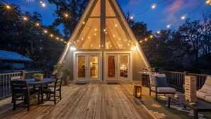 Une terrasse extérieure accueillante, dotée d’un plancher en bois, d’une table à manger avec des chaises et d’un coin salon avec un fauteuil rembourré et une petite table.