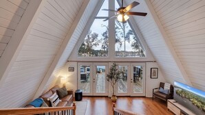 Un loft espaiós amb un sostre alt, un ventilador de sostre, un televisor gran i vista als arbres a través de les finestres.