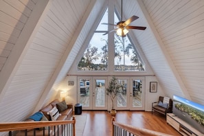 Un loft espacioso con techos altos, un ventilador de techo, un televisor grande y vistas a árboles a través de las ventanas.