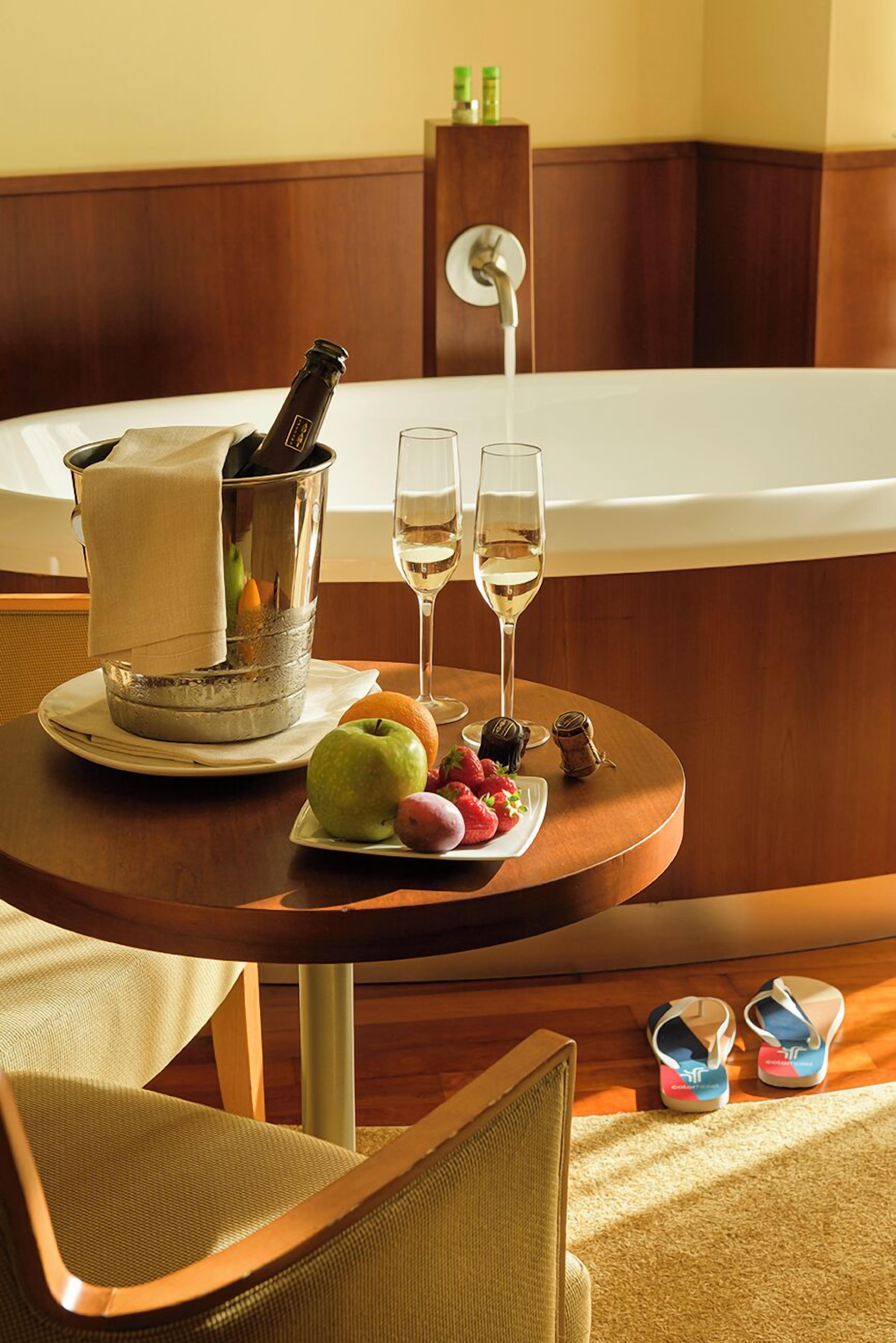 A hotel room with a round wooden table set with fruit, a bottle of champagne, and two glasses of wine.