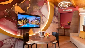 Moderni hotellihuone, jossa on seinälle asennettu televisio, pyöreä ruokapöytä tuoleineen, sohva ja koristeellinen seinä, jossa on abstraktia taidetta.