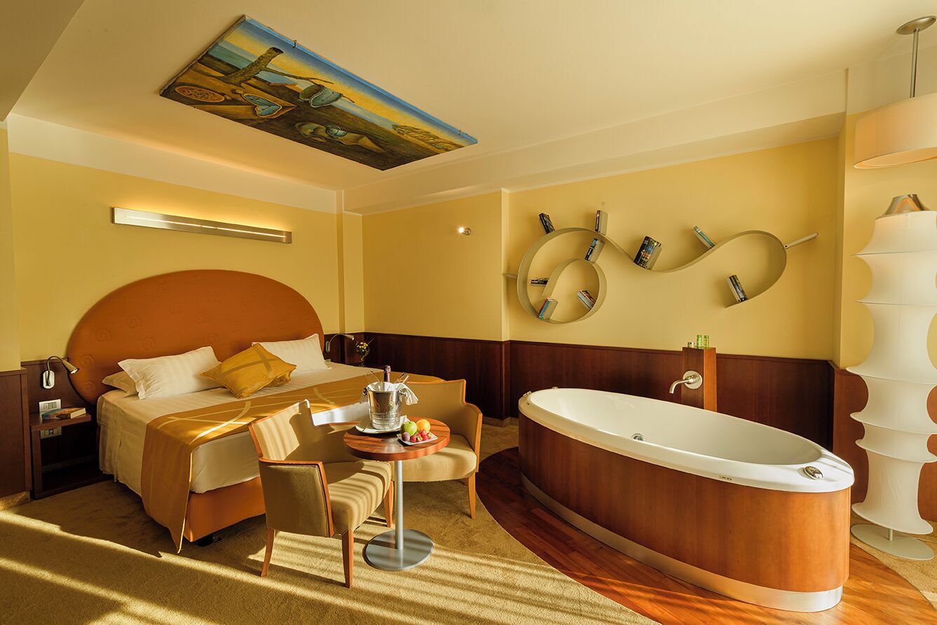 A hotel room with a large bed, a sitting area with a table and chairs, a bathtub, and a wall-mounted mirror with books.