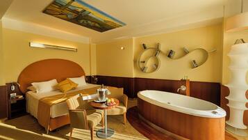 A hotel room with a large bed, a sitting area with a table and chairs, a bathtub, and a wall-mounted mirror with books.