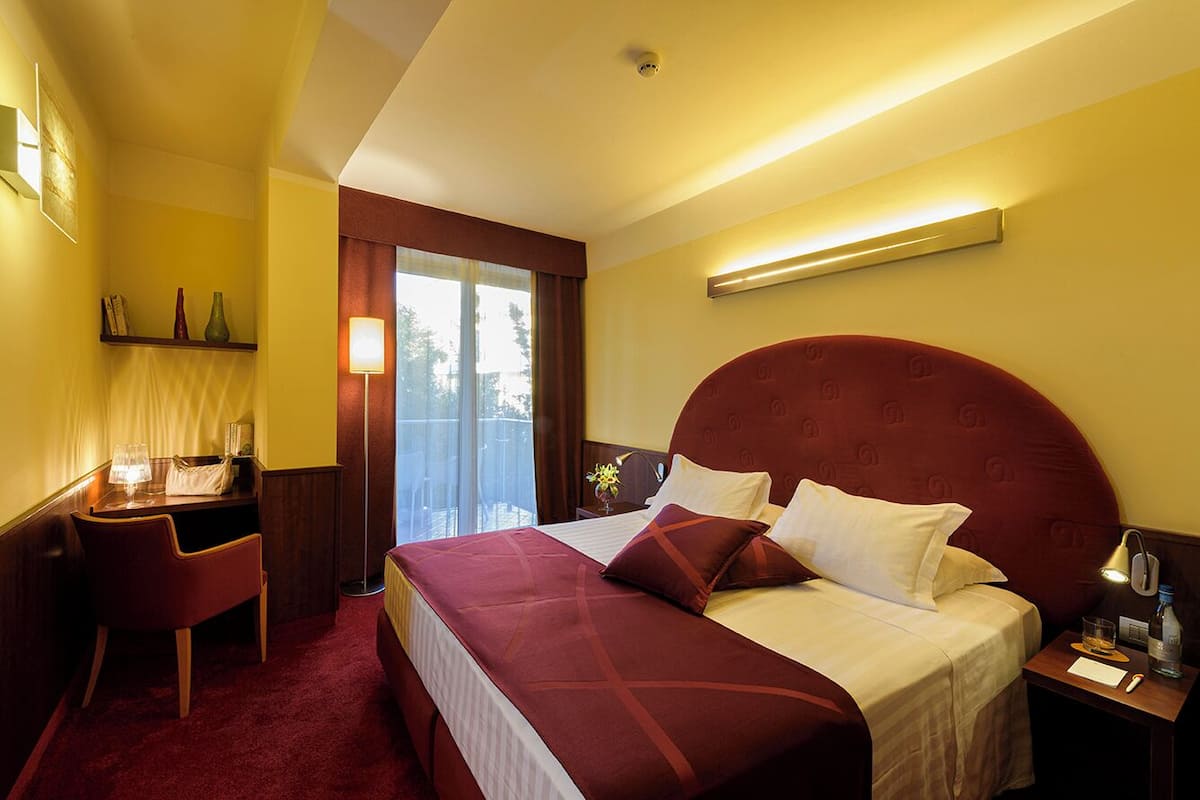 Ein Hotelzimmer mit einem großen Bett, einem Schreibtisch mit Stuhl, einem Nachttisch mit Lampe und einem Fenster mit Vorhängen.