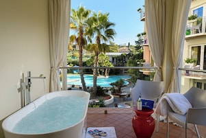 Un balcón con una bañera, una mesa roja y vistas a una piscina y palmeras.