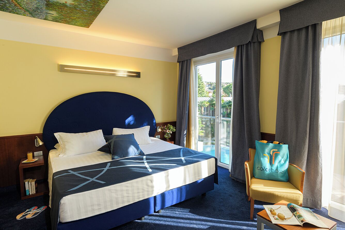 Habitación de hotel con una cama grande, una silla, una mesa con material de lectura y vistas al exterior.