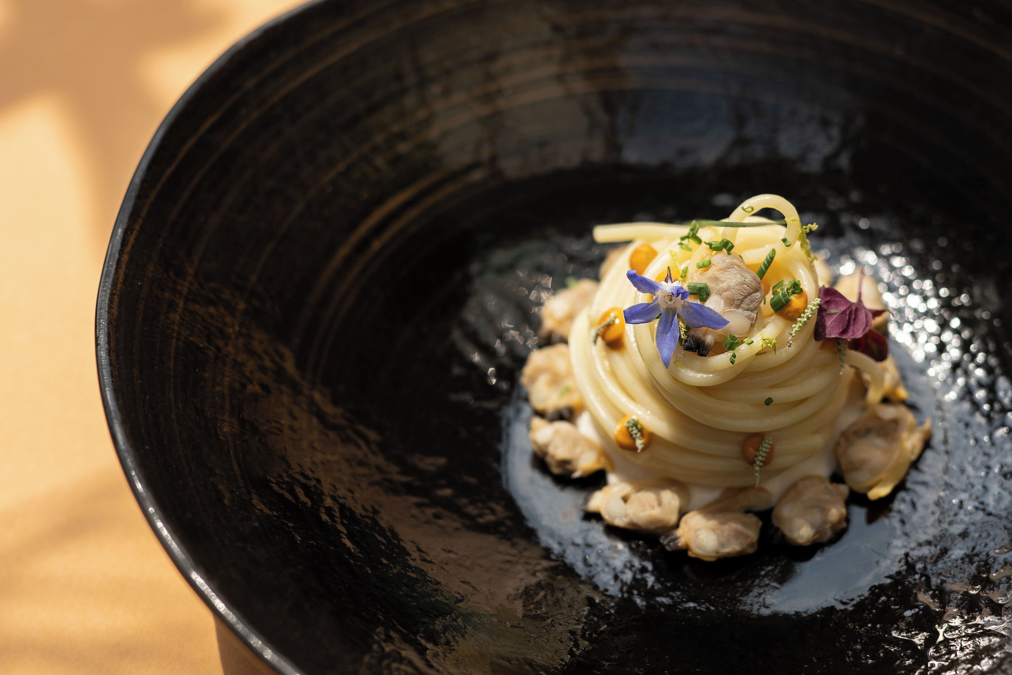 A black bowl containing a layered dish with a central spiral, garnished with small flowers and herbs.