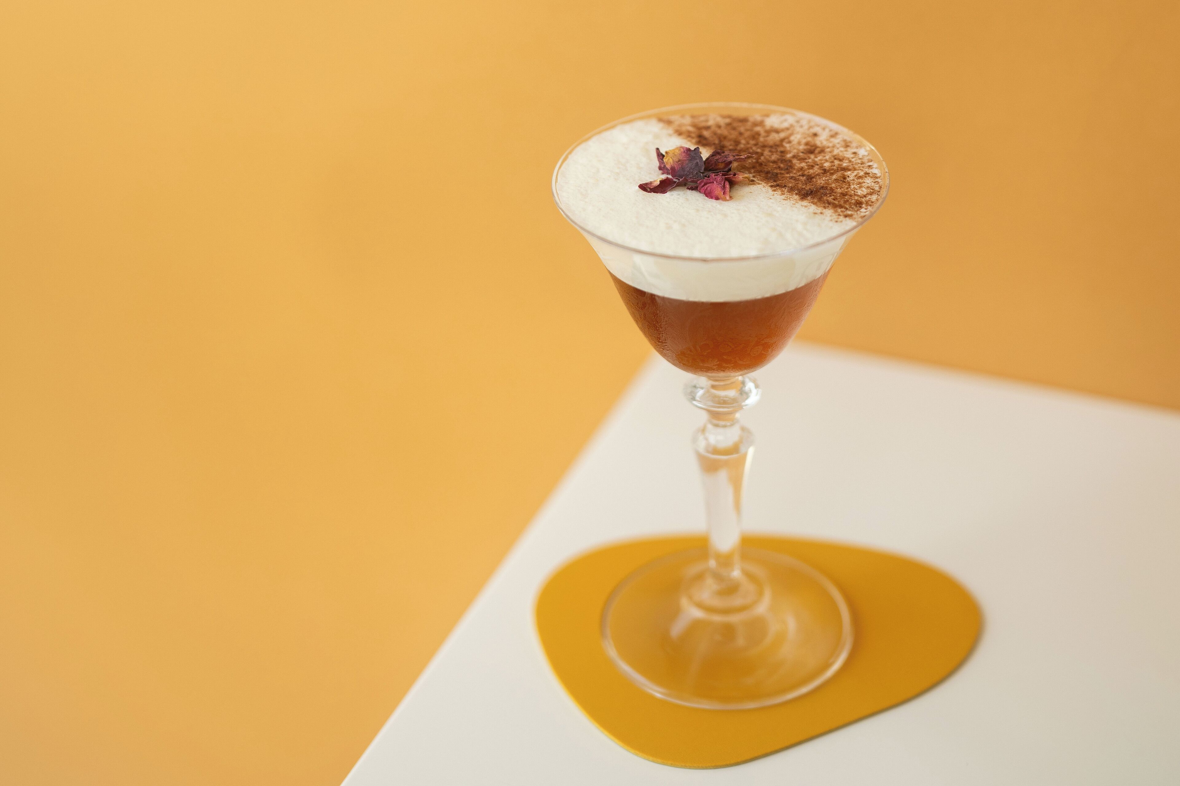 Una copa de martini con una bebida en capas, una guarnición de flor y un toque de cacao espolvoreado encima, sobre un fondo amarillo.