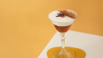 Una copa de martini con una bebida en capas, una guarnición de flor y un toque de cacao espolvoreado encima, sobre un fondo amarillo.