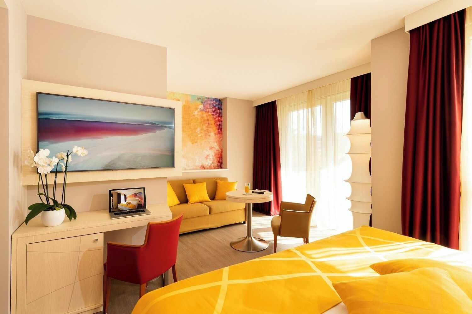 A hotel room with a bed, a desk, a sofa, a chair, a TV, and a painting.