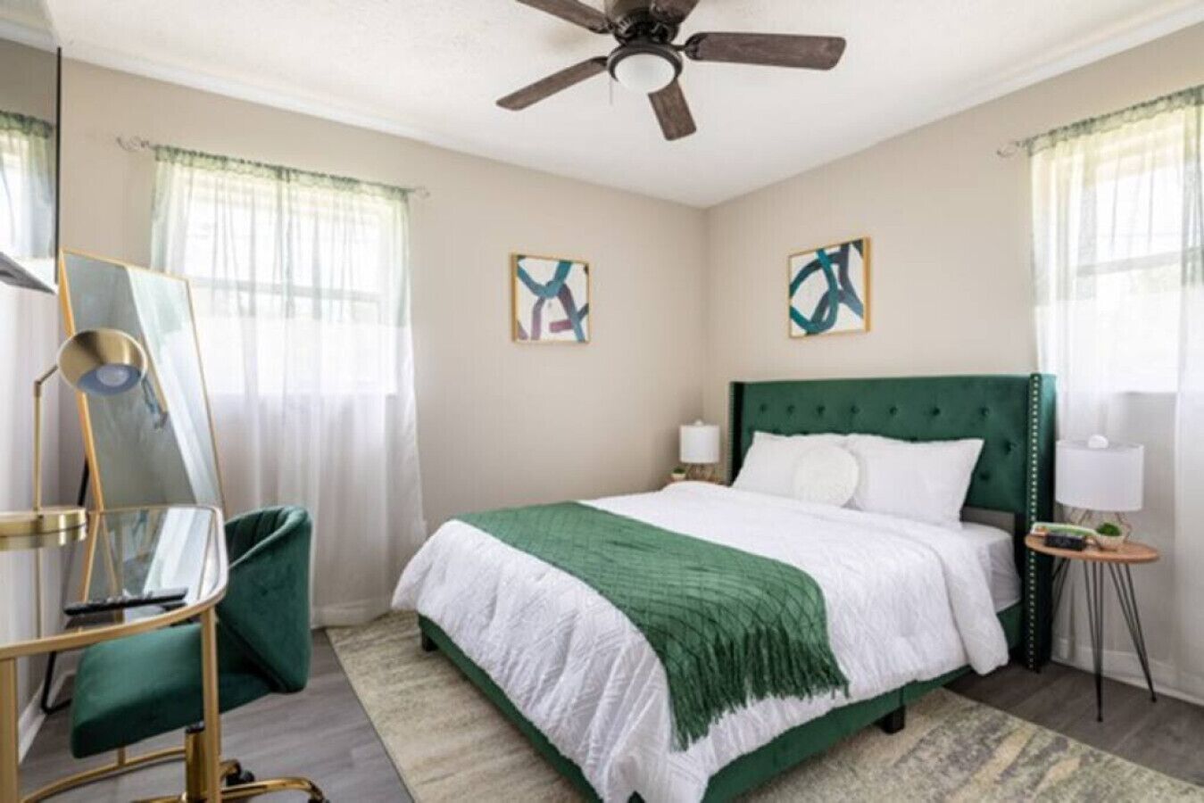 Une chambre d’hôtel avec un lit recouvert d’un tissu vert, un ventilateur de plafond, deux tableaux accrochés aux murs, une table d’appoint avec une lampe et un fauteuil vert.