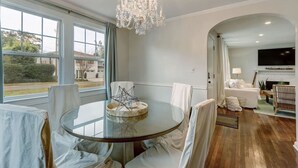 A spacious living room with a round glass table, white chairs, a chandelier, and a fireplace.
