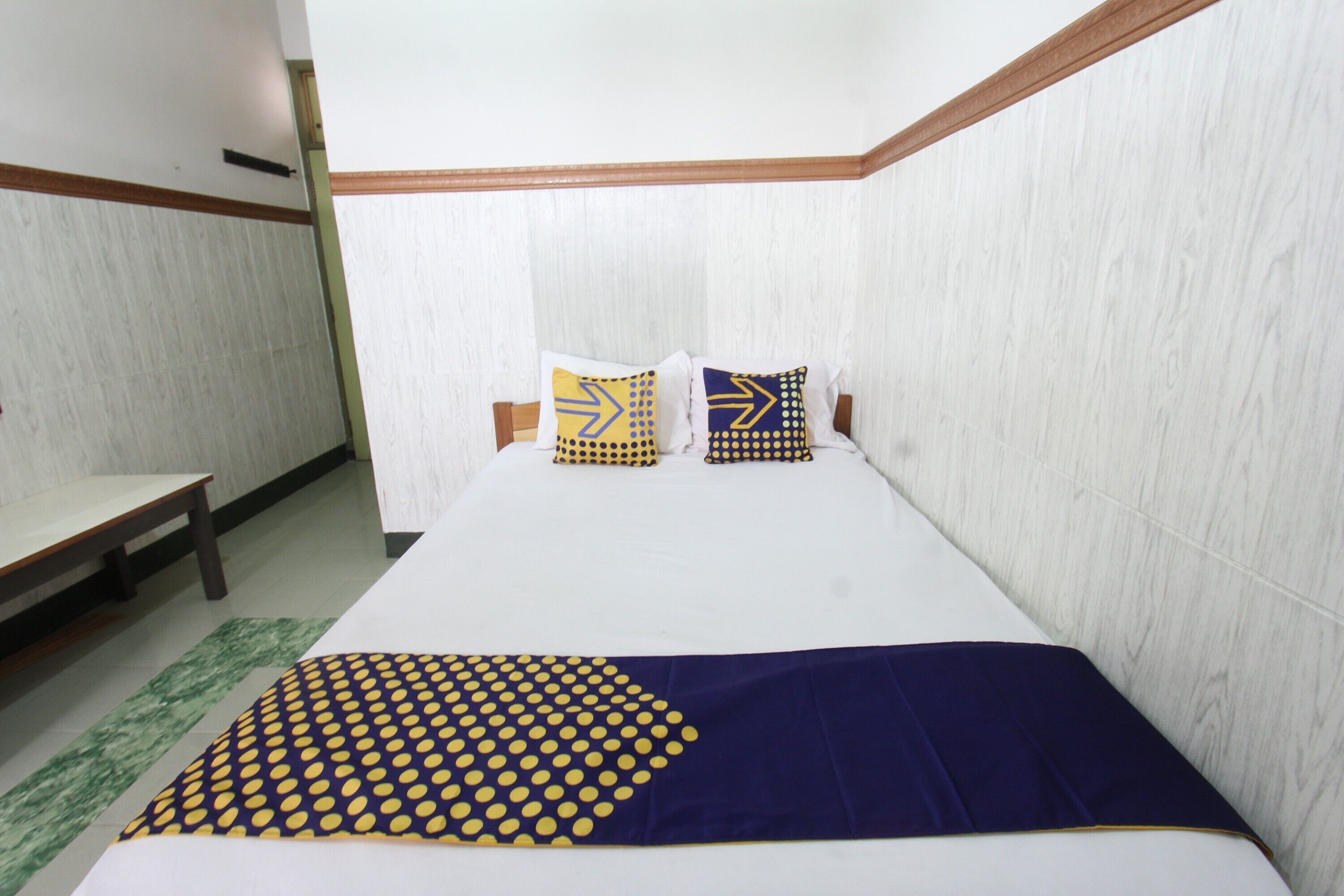 A hotel room with a large bed, two pillows with geometric patterns, a wooden bench, and a green rug.