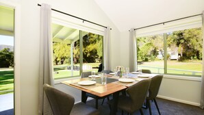 Un coin repas avec une table dressée pour quatre personnes, de grandes fenêtres avec des rideaux et une vue sur un espace extérieur verdoyant.