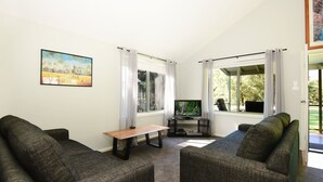 Un salon chaleureux avec deux canapés gris, une table basse en bois et un téléviseur posé sur un meuble. De grandes fenêtres sont ornées de rideaux, un tableau est accroché au mur et une porte donne sur l’extérieur.