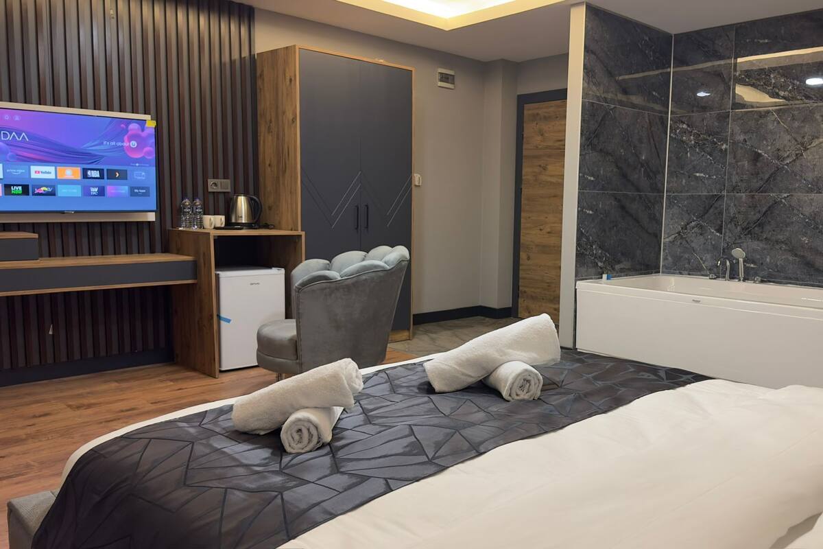 Une chambre d’hôtel moderne dotée d’un grand lit, d’un téléviseur intégré, d’un petit réfrigérateur, d’un coin salon et d’une grande salle de bain avec une baignoire et des murs en marbre.