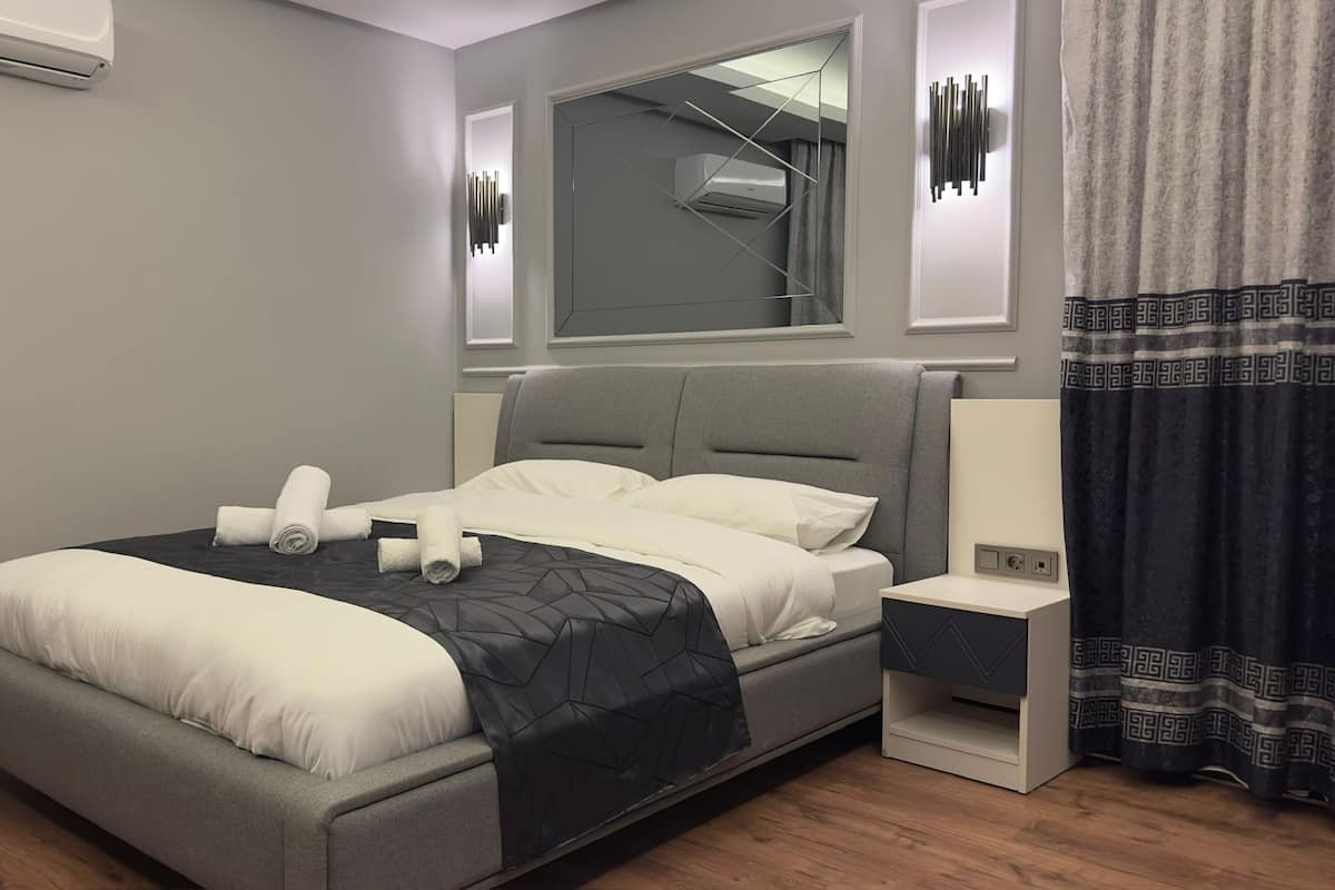 Une chambre d’hôtel moderne avec un lit gris, du linge de lit blanc, une table de chevet avec une lampe et un grand miroir au-dessus du lit.
