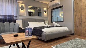 A modern hotel room with a grey sofa, a wooden table, a bed with a grey headboard, and a large window with blinds. - Wonne Boutique Hotel (Ankara)