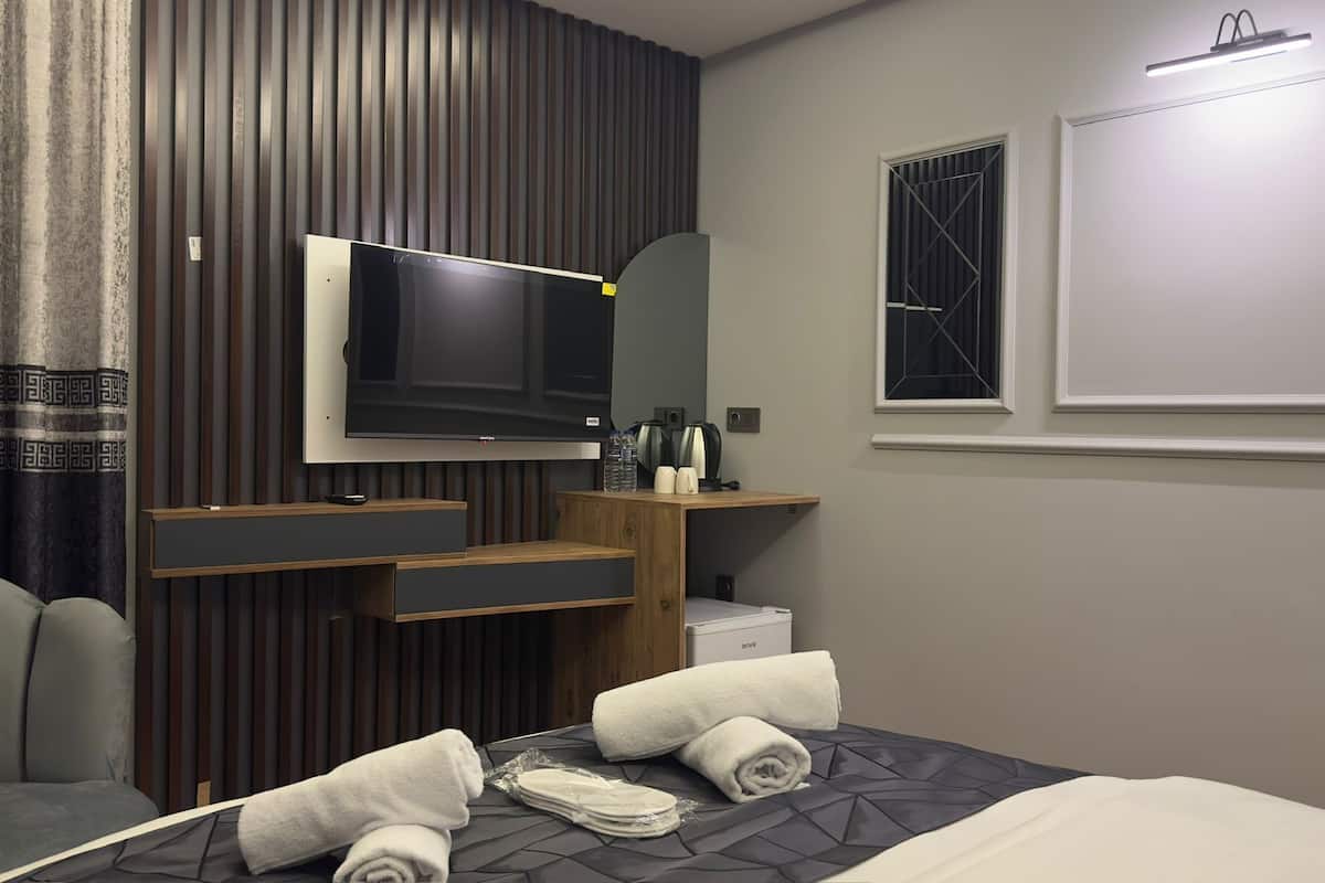 Une chambre d’hôtel moderne dotée d’un grand téléviseur à écran plat fixé au mur, d’une coiffeuse en bois avec un miroir, d’un petit lavabo et d’un lit avec des serviettes soigneusement pliées.