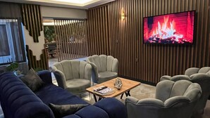 A modern living room with a large flat-screen TV displaying a fireplace, a dark blue sofa, and grey armchairs. - Wonne Boutique Hotel (Ankara)