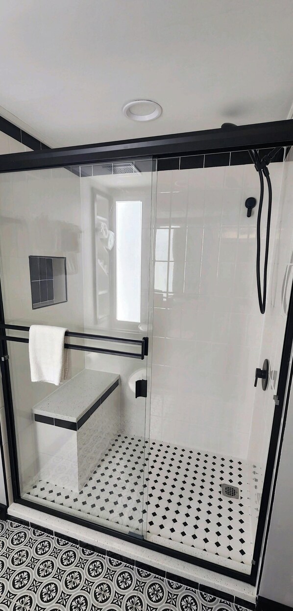 Una ducha con paredes de cristal, suelo de baldosas blancas y un borde decorativo con mosaicos blancos y negros.
